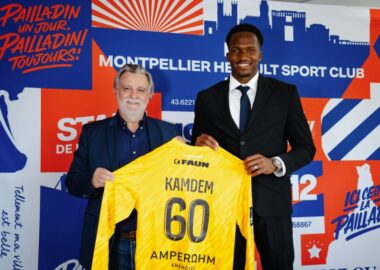 Two men in smart-casual attire hold up a bright yellow sports jersey labeled Kamdem 60 in front of a colorful club backdrop with logos and graphics.