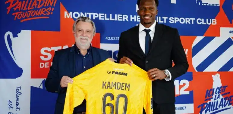 Two men in smart-casual attire hold up a bright yellow sports jersey labeled Kamdem 60 in front of a colorful club backdrop with logos and graphics.