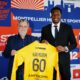 Two men in smart-casual attire hold up a bright yellow sports jersey labeled Kamdem 60 in front of a colorful club backdrop with logos and graphics.