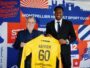 Two men in smart-casual attire hold up a bright yellow sports jersey labeled Kamdem 60 in front of a colorful club backdrop with logos and graphics.