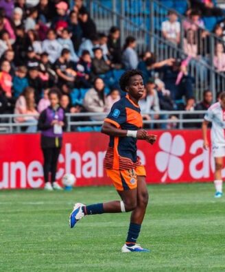 Soccer player in orange and navy kit running on a green field during a match, with spectators in the stands behind.