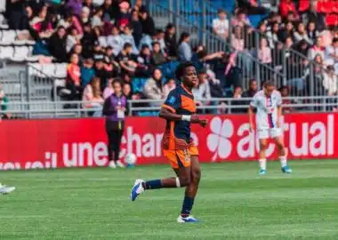 Soccer player in orange and navy kit running on a green field during a match, with spectators in the stands behind.