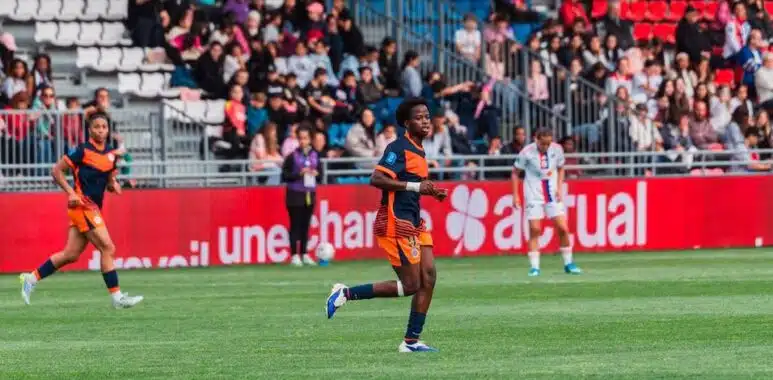 Soccer player in orange and navy kit running on a green field during a match, with spectators in the stands behind.