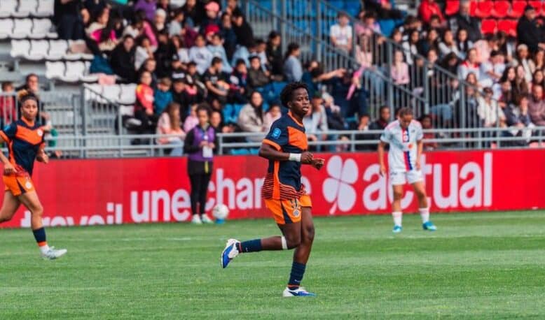 Soccer player in orange and navy kit running on a green field during a match, with spectators in the stands behind.