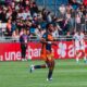 Soccer player in orange and navy kit running on a green field during a match, with spectators in the stands behind.