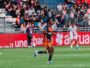 Soccer player in orange and navy kit running on a green field during a match, with spectators in the stands behind.