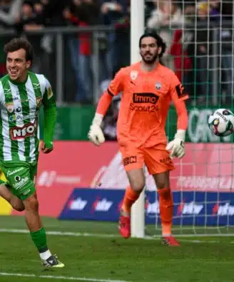 Soccer scene: a green-and-white striped player celebrates near the goal as an orange-clad goalkeeper watches the ball in the air by the net.