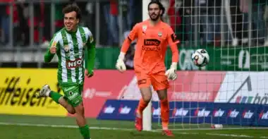 Sacha Delaye Lustenau | AllezPaillade.com Soccer scene: a green-and-white striped player celebrates near the goal as an orange-clad goalkeeper watches the ball in the air by the net.