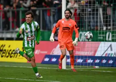 Soccer scene: a green-and-white striped player celebrates near the goal as an orange-clad goalkeeper watches the ball in the air by the net.