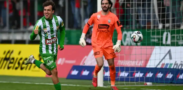 Sacha Delaye Lustenau | AllezPaillade.com Soccer scene: a green-and-white striped player celebrates near the goal as an orange-clad goalkeeper watches the ball in the air by the net.