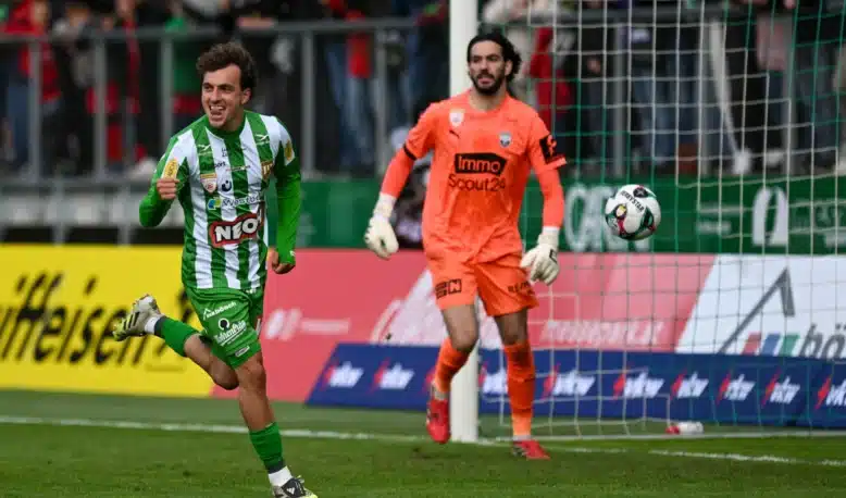 Sacha Delaye Lustenau | AllezPaillade.com Soccer scene: a green-and-white striped player celebrates near the goal as an orange-clad goalkeeper watches the ball in the air by the net.
