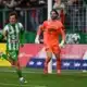 Soccer scene: a green-and-white striped player celebrates near the goal as an orange-clad goalkeeper watches the ball in the air by the net.