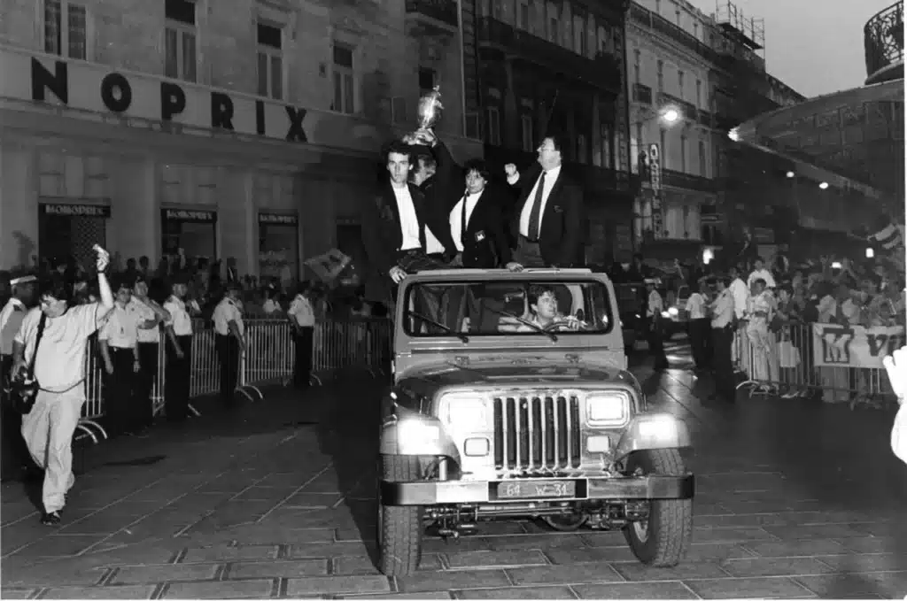 Three men in tuxedos stand on the hood and roof of a jeep, leading a nighttime parade as the crowd cheers along a lined street with barricades and storefronts behind them.