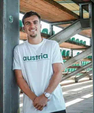filtersquality80 | AllezPaillade.com Man leaning against a metal pillar in an outdoor stadium structure, wearing a white shirt that says 'austria' with green seats visible in the background