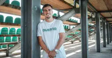 Man leaning against a metal pillar in an outdoor stadium structure, wearing a white shirt that says 'austria' with green seats visible in the background