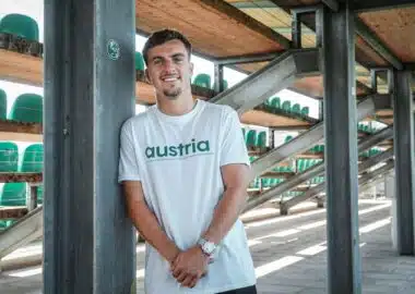 Man leaning against a metal pillar in an outdoor stadium structure, wearing a white shirt that says 'austria' with green seats visible in the background