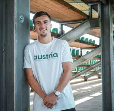Man leaning against a metal pillar in an outdoor stadium structure, wearing a white shirt that says 'austria' with green seats visible in the background