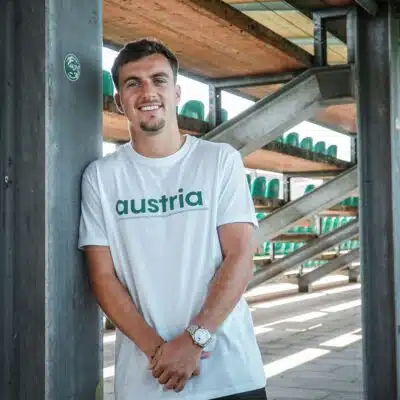 Man leaning against a metal pillar in an outdoor stadium structure, wearing a white shirt that says 'austria' with green seats visible in the background