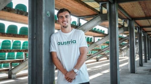 Man leaning against a metal pillar in an outdoor stadium structure, wearing a white shirt that says 'austria' with green seats visible in the background
