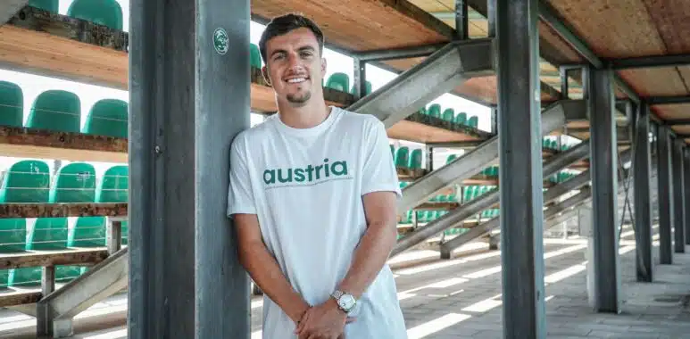 filtersquality80 | AllezPaillade.com Man leaning against a metal pillar in an outdoor stadium structure, wearing a white shirt that says 'austria' with green seats visible in the background