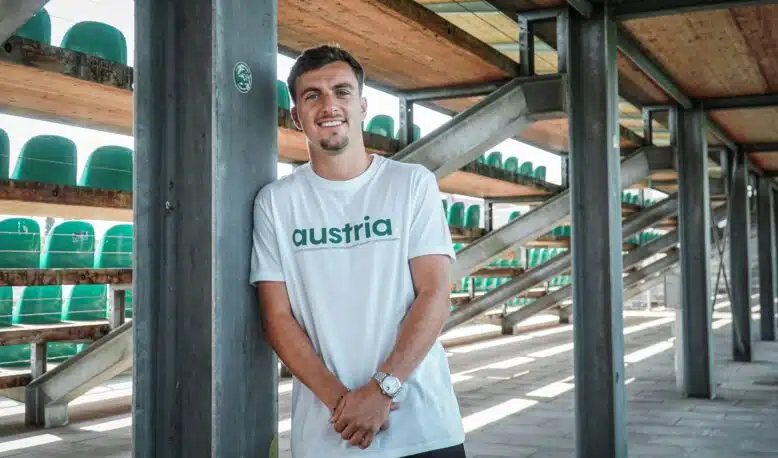 Man leaning against a metal pillar in an outdoor stadium structure, wearing a white shirt that says 'austria' with green seats visible in the background