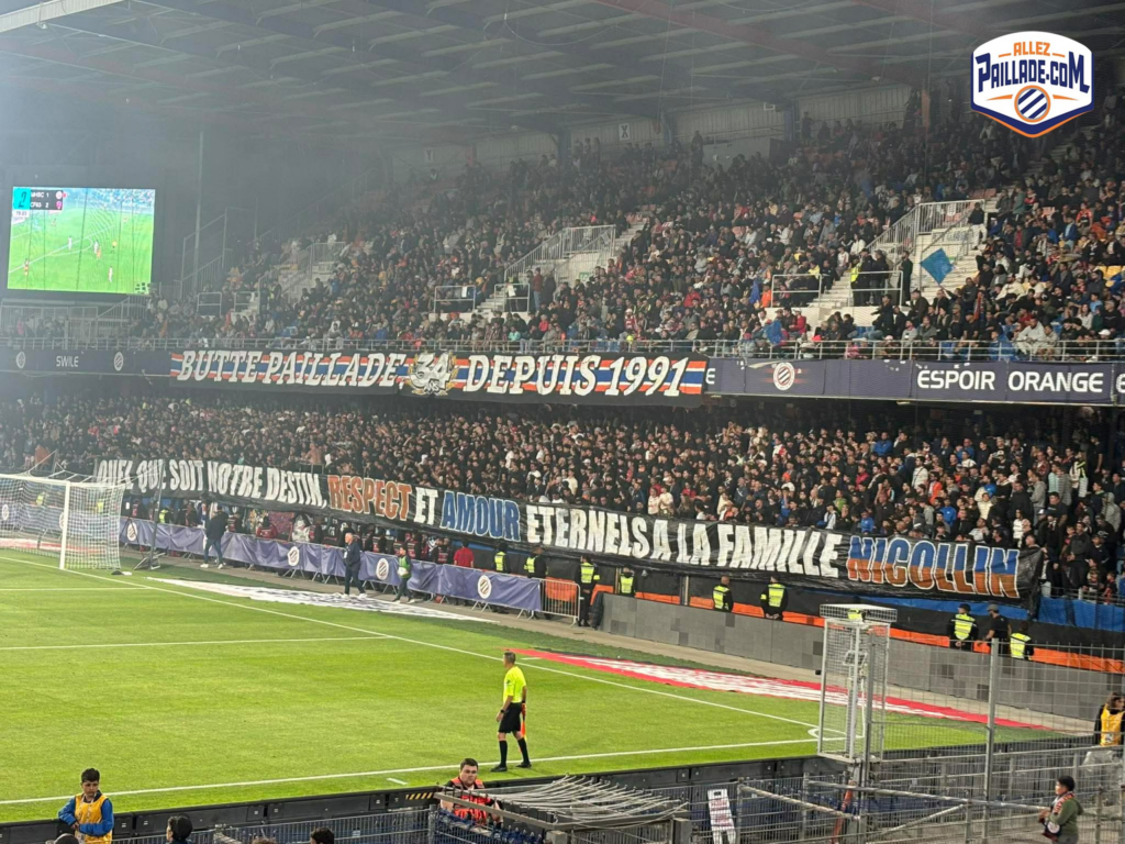 Packed soccer stadium with banners and fans in the stands; a large video screen is on the left.