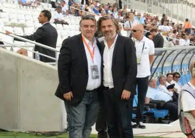 Two men in dark blazers pose for a photo at a football stadium, with spectators in the stands behind them and media around.