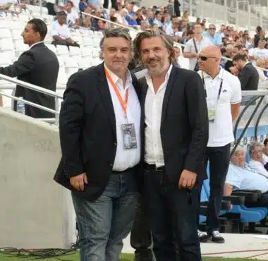Two men in dark blazers pose for a photo at a football stadium, with spectators in the stands behind them and media around.