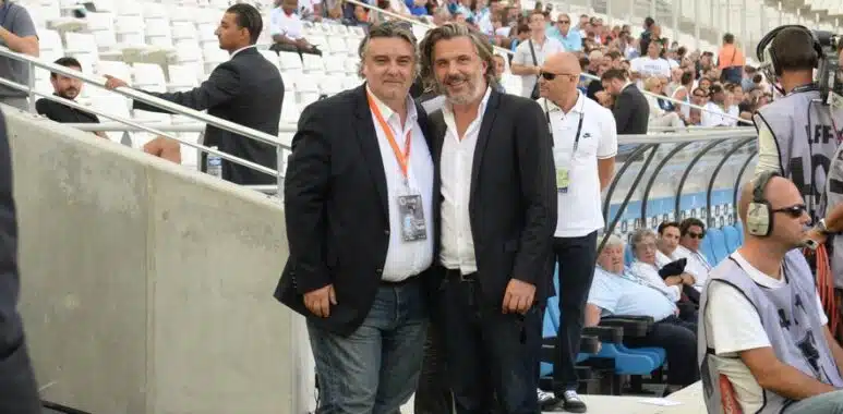 Two men in dark blazers pose for a photo at a football stadium, with spectators in the stands behind them and media around.