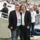 Two men in dark blazers pose for a photo at a football stadium, with spectators in the stands behind them and media around.