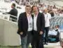 Two men in dark blazers pose for a photo at a football stadium, with spectators in the stands behind them and media around.