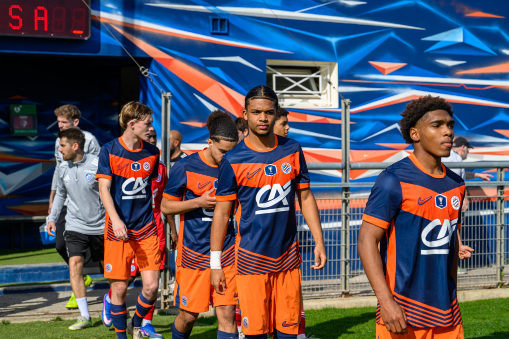 Soccer players in blue and orange uniforms walking out onto the field from a tunnel, with a colorful backdrop behind them.