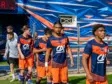 Soccer players in blue and orange uniforms walking out onto the field from a tunnel, with a colorful backdrop behind them.