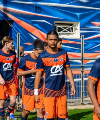 Soccer players in blue and orange uniforms walking out onto the field from a tunnel, with a colorful backdrop behind them.