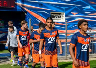 Soccer players in blue and orange uniforms walking out onto the field from a tunnel, with a colorful backdrop behind them.
