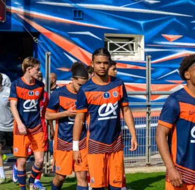Soccer players in blue and orange uniforms walking out onto the field from a tunnel, with a colorful backdrop behind them.