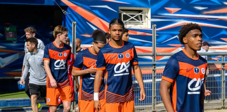 Soccer players in blue and orange uniforms walking out onto the field from a tunnel, with a colorful backdrop behind them.