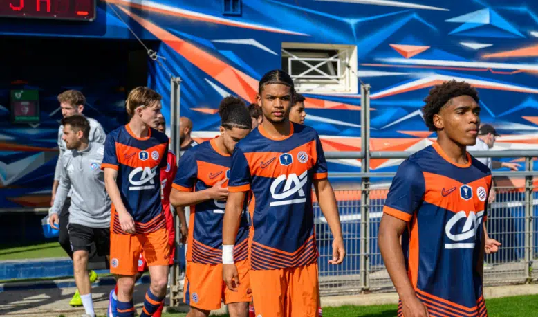 Soccer players in blue and orange uniforms walking out onto the field from a tunnel, with a colorful backdrop behind them.