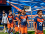 Soccer players in blue and orange uniforms walking out onto the field from a tunnel, with a colorful backdrop behind them.