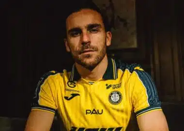 Man with short dark hair and stubble wearing a yellow PAU football jersey, seated in a dimly lit room.