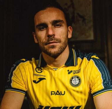 Man with short dark hair and stubble wearing a yellow PAU football jersey, seated in a dimly lit room.