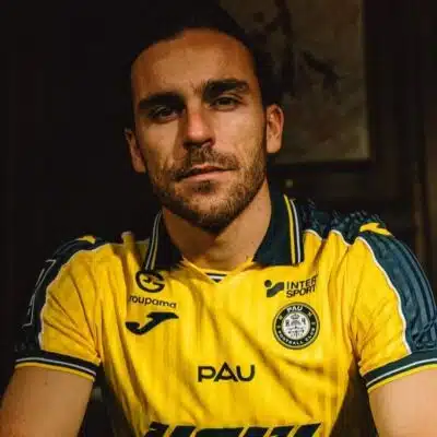 Man with short dark hair and stubble wearing a yellow PAU football jersey, seated in a dimly lit room.