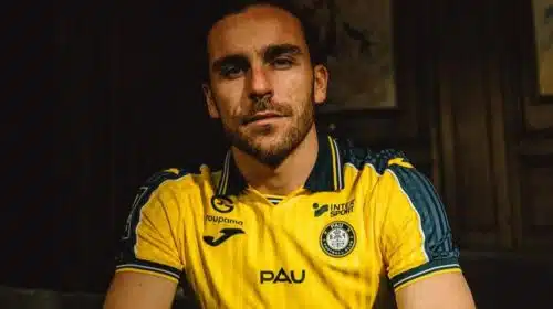Man with short dark hair and stubble wearing a yellow PAU football jersey, seated in a dimly lit room.
