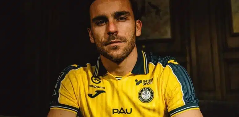 Man with short dark hair and stubble wearing a yellow PAU football jersey, seated in a dimly lit room.