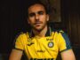 Man with short dark hair and stubble wearing a yellow PAU football jersey, seated in a dimly lit room.