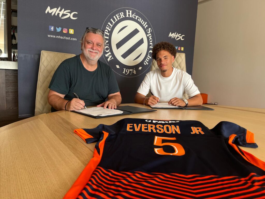 Everson Laurent Nicollin | AllezPaillade.com Two men sign documents at a table in a room with a club banner behind them; a navy-orange Everson Jr. jersey with the number 5 lies on the table in the foreground.