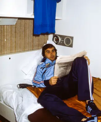 Young man in a blue tracksuit lounging on a bed, reading a newspaper in a small, retro-style room.