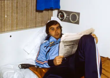 Young man in a blue tracksuit lounging on a bed, reading a newspaper in a small, retro-style room.