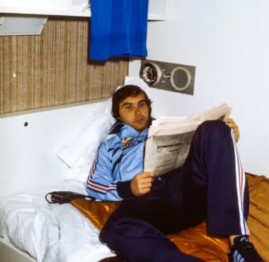 Young man in a blue tracksuit lounging on a bed, reading a newspaper in a small, retro-style room.