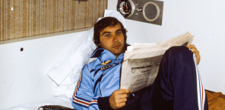 Young man in a blue tracksuit lounging on a bed, reading a newspaper in a small, retro-style room.